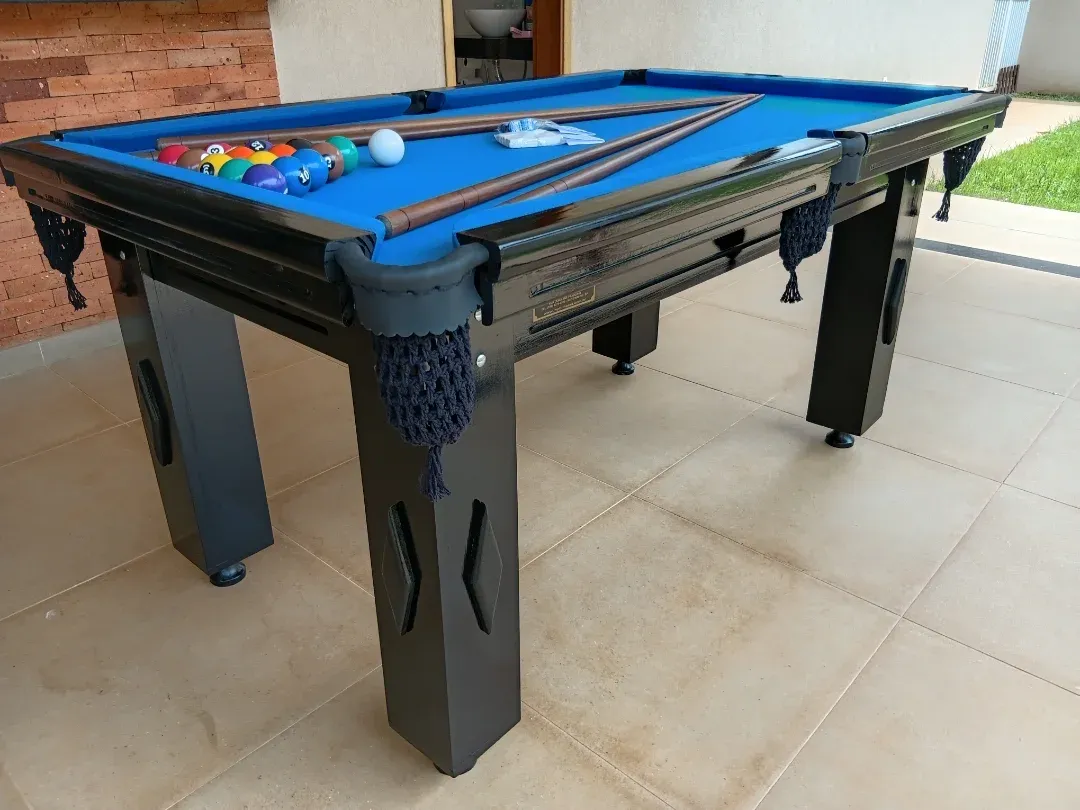 Mesa de bilhar preta com pano azul, bolas e tacos, varanda com gramado ao fundo.