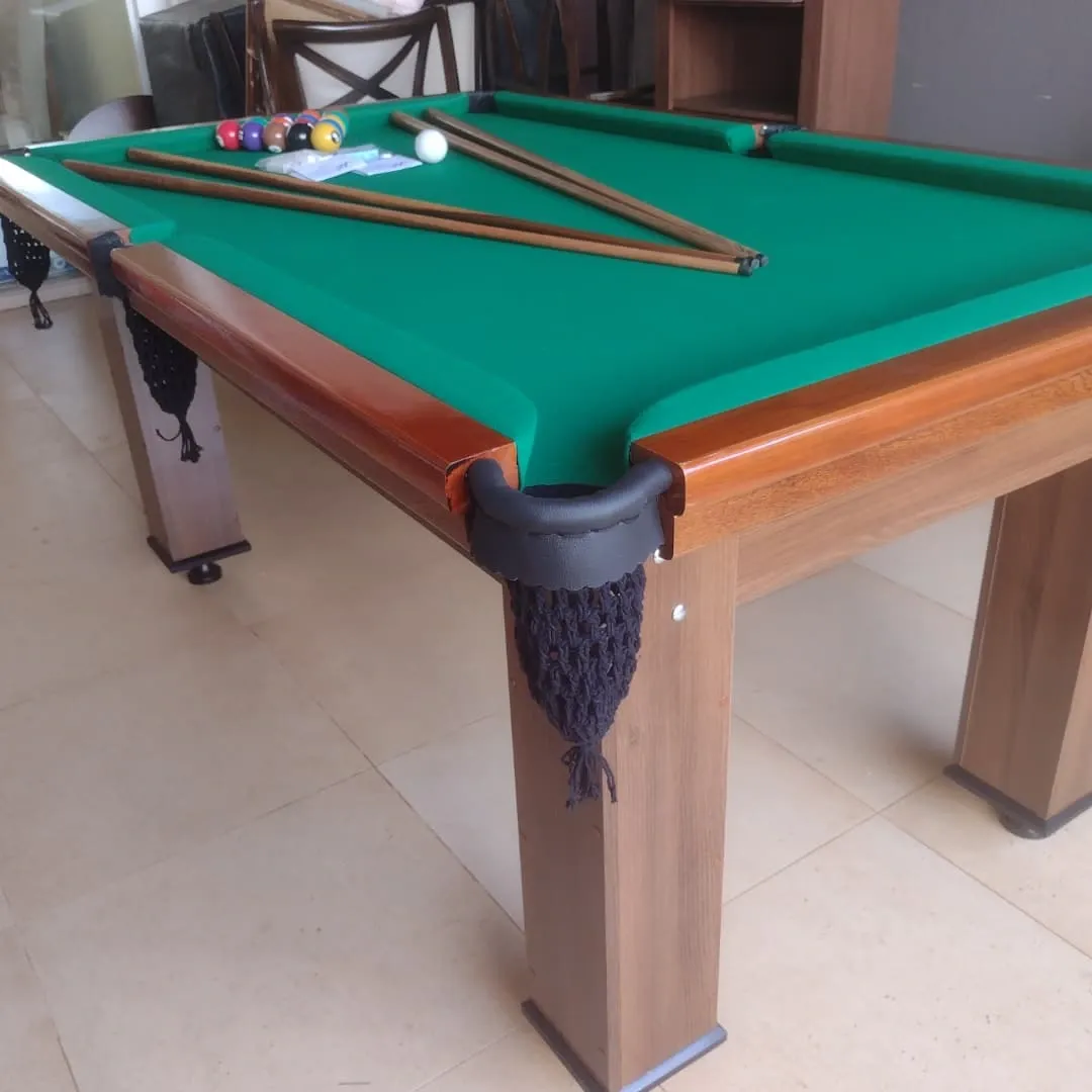 Mesa de bilhar com pano verde, estrutura madeira escura, caçapa com rede e tacos apoiados.